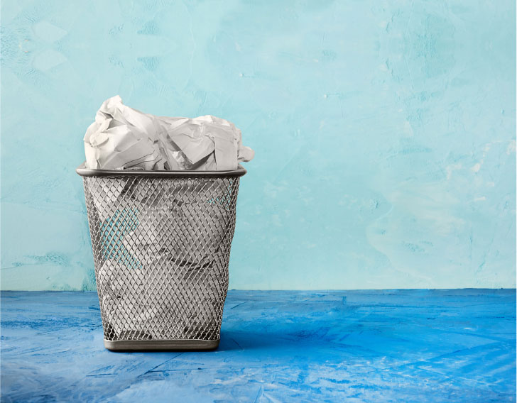 A trash container full of paper on a blue floor in front of a lighter blue wall